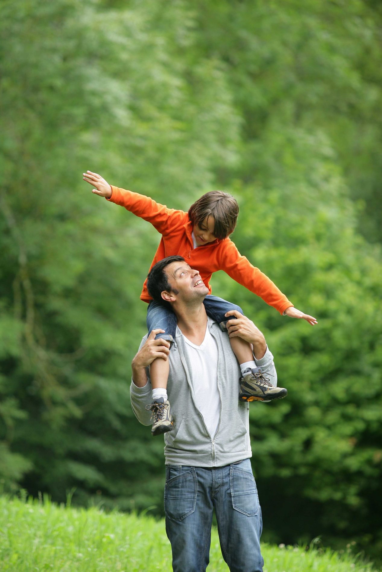 Photo of a man with son on his shoulders outdoors walking through a grassy area.